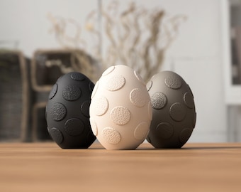 May include: Three decorative egg-shaped objects with a textured surface and large, circular patterns. One is black, one is off-white, and the third is a dark gray. They are arranged on a wooden surface.
