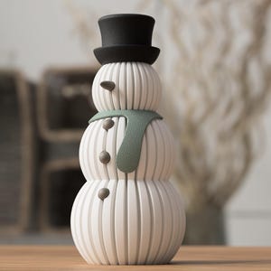 May include: A decorative snowman figurine with a black top hat, a green scarf, and three dark gray buttons. The snowman is white with vertical lines and has two small, dark gray eyes. A festive winter decoration.