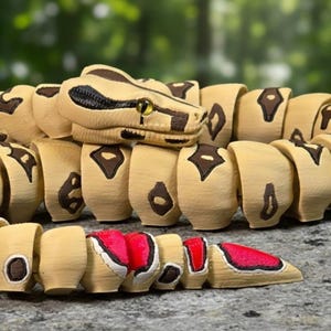 Puede incluir: Un juguete de serpiente articulada de madera con cuerpo beige, marcas marrones y detalles rojos. La cabeza de la serpiente está detallada con patrones negros y marrones y ojos amarillos. El juguete está sobre una superficie de piedra gris.
