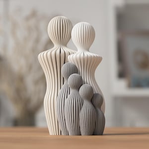 Op de afbeelding: Een houten familiesculptuur met twee volwassen figuren en drie kleinere figuren, allemaal met verticale strepen. De figuren zijn in wit- en grijstinten, geplaatst op een houten oppervlak. Een decoratief object.