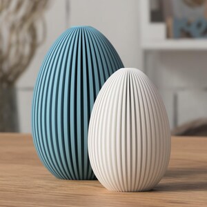 May include: Two decorative egg-shaped objects. One is a light blue color, and the other is white. Both have vertical grooves. They are placed on a wooden surface. The background is blurred, suggesting a home decor setting.