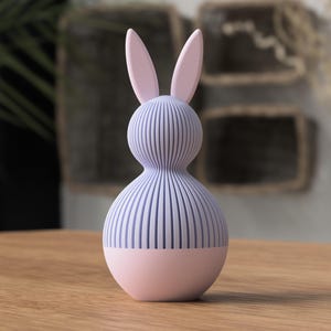 Könnte beinhalten: Eine dekorative Hasenfigur in Pastellfarben. Der Hase hat einen hellvioletten Körper mit vertikalen Linien, rosa Ohren und einen rosa Sockel. Die Figur besteht aus einem glatten Material und steht auf einer Holzoberfläche.