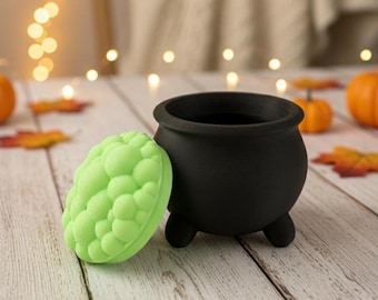 May include: A black cauldron with three small feet and a light green, textured, oval-shaped item next to it. The cauldron and the green item are on a light-colored wooden surface. Small pumpkins and fall leaves are scattered around.