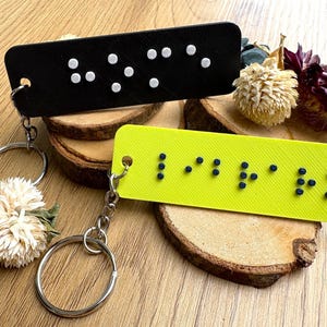 May include: Two rectangular keychains, one black and one yellow, with braille text. The black keychain has white dots, and the yellow keychain spells "I LOVE YOU" in black braille. Both have silver keyrings.