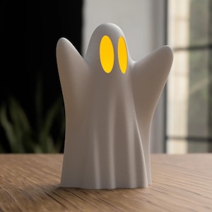 Könnte beinhalten: Eine weiße, geisterförmige dekorative Lampe mit zwei gelben, ovalen Augen. Die Lampe hat ein drapiertes Design und steht auf einer Holzoberfläche. Der Hintergrund ist verschwommen, mit einem dunklen Bereich und einem Fenster.