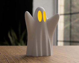May include: A white ghost-shaped decorative lamp with two yellow oval-shaped eyes. The lamp has a draped design and is sitting on a wooden surface. The background is blurred, with a dark area and a window.
