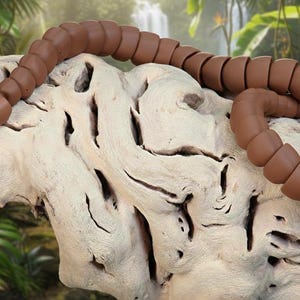Può includere: Un giocattolo a forma di serpente articolato marrone è appoggiato su una superficie di legno chiaro e strutturato. Il serpente è segmentato, consentendo una posa flessibile. Lo sfondo presenta un rigoglioso fogliame verde e una cascata.