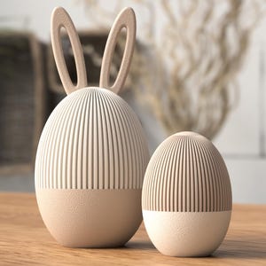 Pode incluir: Dois objetos decorativos em forma de ovo de Páscoa. O maior tem orelhas de coelho. Ambos são creme com linhas verticais na metade superior e uma base texturizada e sólida. Estão sobre uma superfície de madeira.