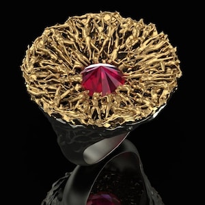 May include: A gold ring featuring a large red gemstone surrounded by a ring of tiny gold figures. The ring is set against a black background.