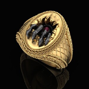 Könnte beinhalten: Ein goldener Ring mit einem detaillierten Design, das eine schwarze Klaue zeigt, die aus einem Loch in der Mitte herausragt. Die Klaue hat einen roten Edelstein in der Mitte.