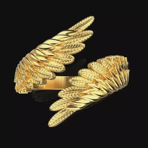 Puede incluir: Un anillo dorado con diseño de alas. El anillo presenta dos alas detalladas que se envuelven alrededor del dedo. Las alas están hechas de plumas individuales, creando una apariencia texturizada. El anillo está sobre un fondo negro.