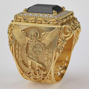 Puede incluir: Anillo dorado con una gran piedra negra rectangular. El anillo presenta detalles intrincados, incluyendo un &aacute;ngel con alas y texto religioso. Peque&ntilde;as piedras claras rodean la piedra negra. Una pieza de joyer&iacute;a llamativa.
