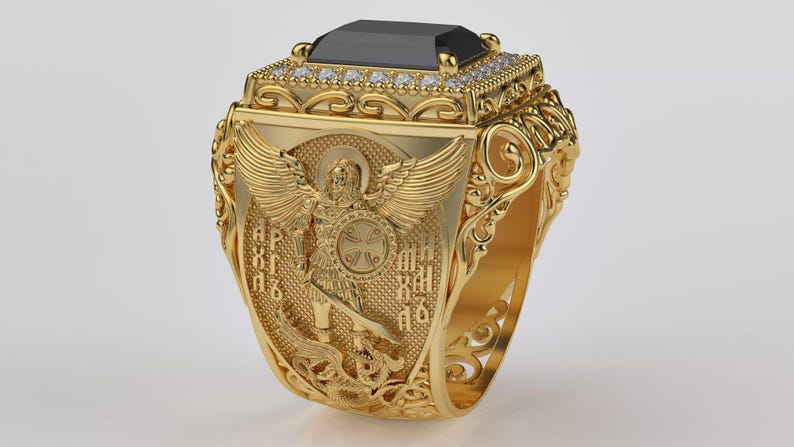 Puede incluir: Anillo dorado con una gran piedra negra rectangular. El anillo presenta detalles intrincados, incluyendo un &aacute;ngel con alas y texto religioso. Peque&ntilde;as piedras claras rodean la piedra negra. Una pieza de joyer&iacute;a llamativa.