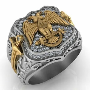 May include: A detailed silver ring with gold accents, featuring an eagle emblem with a triangle and the text "DEUS MEUMQUE JUS". The ring is encrusted with small, clear gemstones around the emblem.