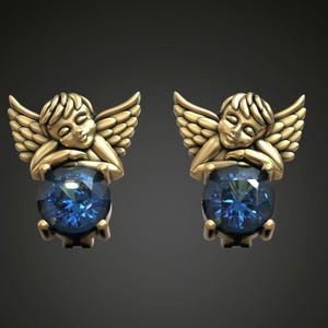 May include: A pair of gold-colored angel earrings. Each earring features a sleeping angel with outstretched wings, cradling a large, faceted, blue gemstone. The earrings are set against a dark background.