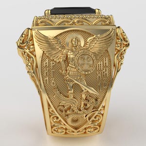 Puede incluir: Un anillo dorado con un dise&ntilde;o ornamentado. El anillo presenta un escudo central con un &aacute;ngel sosteniendo una espada y un escudo. El anillo tiene una piedra negra rectangular en la parte superior y est&aacute; muy detallado con volutas.