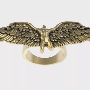 Puede incluir: Un anillo dorado con una escultura detallada de una figura con alas extendidas. El diseño del anillo incorpora una forma humana con alas emplumadas, creando una pieza de joyería única y artística.