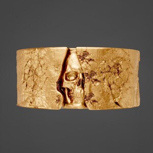 May include: A gold-colored, textured cuff bracelet featuring a skull design. The skull is centered, with the bracelet's sides appearing to split open to reveal it. The bracelet has a rough, cracked texture.