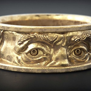 Könnte beinhalten: Ein goldener Ring mit einem detaillierten Design eines Gesichts mit zwei Augen. Der Ring ist aus Metall gefertigt und hat eine glatte, polierte Oberfläche.