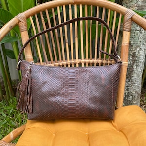 May include: A brown leather handbag with a snakeskin pattern and a long tassel. The bag has a shoulder strap and is sitting on a yellow cushion.