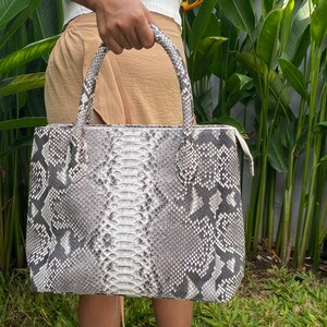 May include: A grey and white snakeskin tote bag with a zip closure. The bag has two top handles.