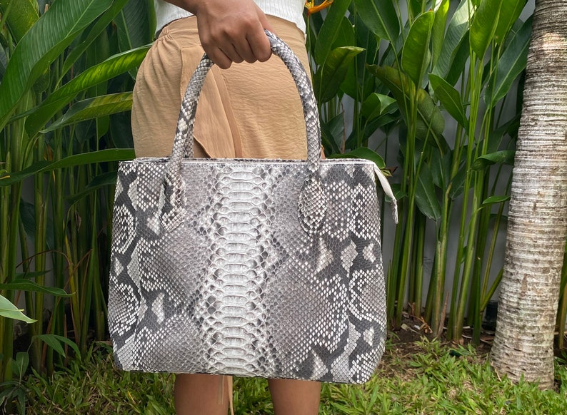 May include: A grey and white snakeskin tote bag with a zip closure. The bag has two top handles.