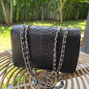以下が含まれることがあります： シルバーのチェーンストラップが付いた黒い革製のハンドバッグ。バッグは、蛇革のような質感の模様をしています。