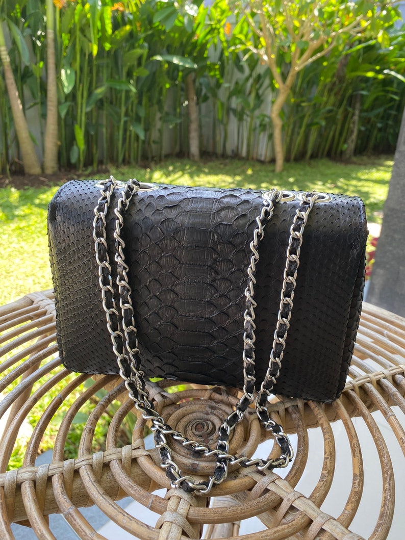 以下が含まれることがあります： シルバーのチェーンストラップが付いた黒い革製のハンドバッグ。バッグは、蛇革のような質感の模様をしています。