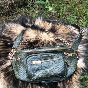 Peut inclure: Une banane en cuir de serpent vert avec une bandoulière en chaîne dorée. Le sac a plusieurs compartiments zippés.