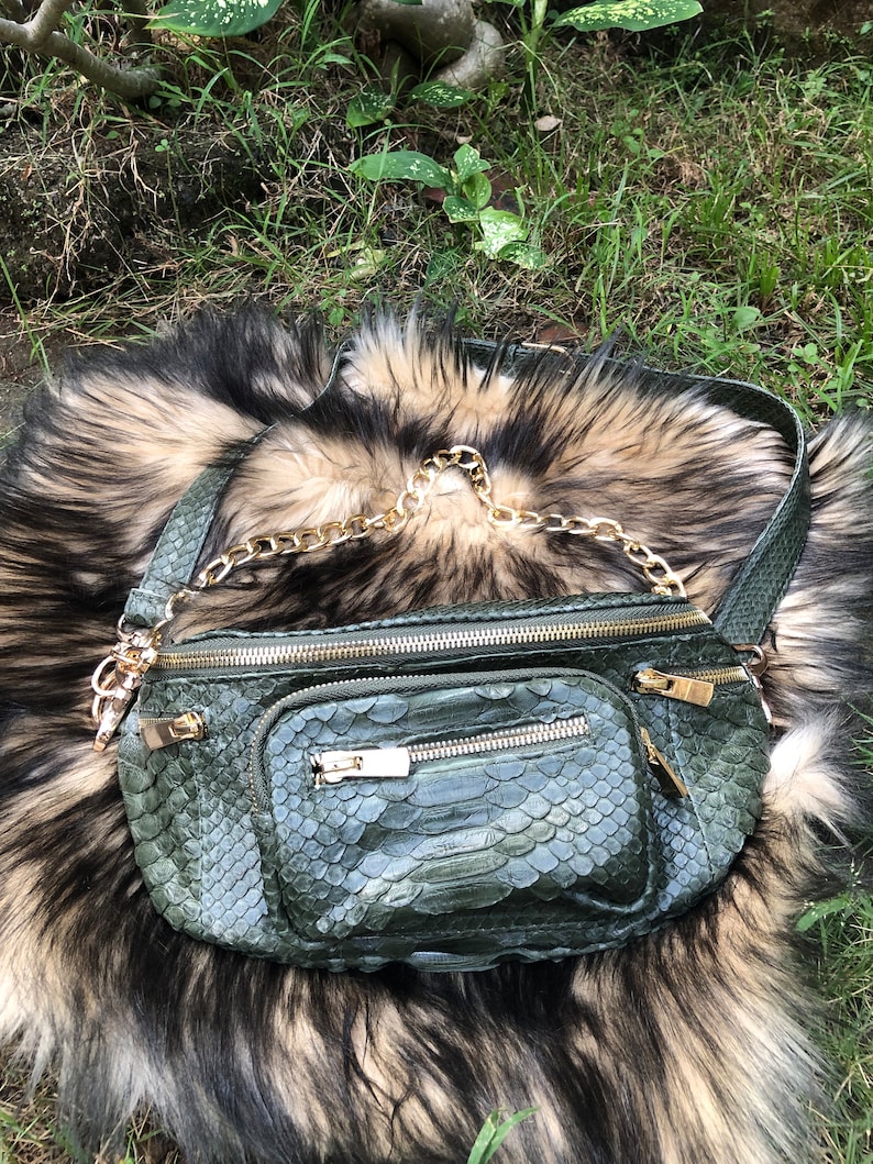 Puede incluir: Una ri&ntilde;onera de piel de serpiente verde con una correa de cadena dorada. La bolsa tiene varios compartimentos con cremallera.