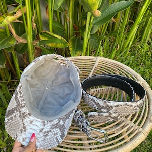 May include: A gray and white snakeskin baseball cap and a matching snakeskin belt with a silver buckle.