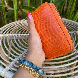 May include: A bright orange leather wallet with a zipper closure. The wallet has a textured, crocodile-like pattern. The wallet is held in a hand with a blue and clear beaded bracelet.
