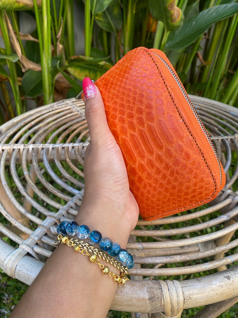 May include: A bright orange leather wallet with a zipper closure. The wallet has a textured, crocodile-like pattern. The wallet is held in a hand with a blue and clear beaded bracelet.