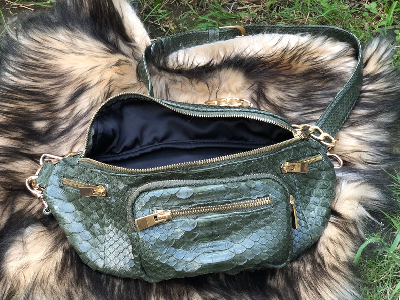 Puede incluir: Una ri&ntilde;onera de piel de serpiente verde con herrajes dorados y una correa de cadena. La bolsa tiene varios compartimentos con cremallera.