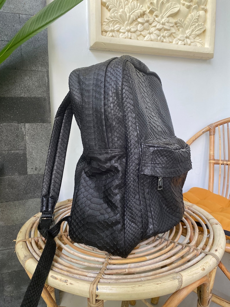 May include: A black leather backpack with a snakeskin pattern. The backpack has a zipped pocket on the front and adjustable straps.