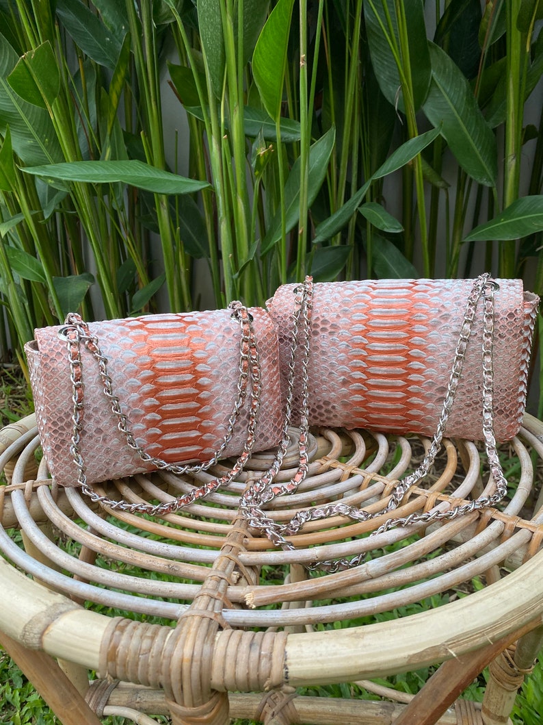 May include: Two small, orange and white snakeskin purses with silver chain straps. The purses are sitting on a woven wicker stool.