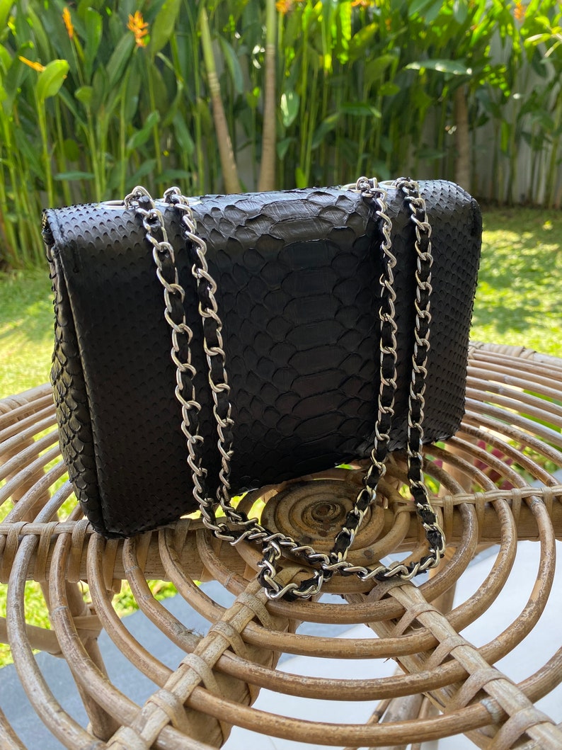 以下が含まれることがあります： 銀色のチェーンストラップが付いた黒いヘビ革のショルダーバッグ。バッグは籐製の椅子の上に置かれています。