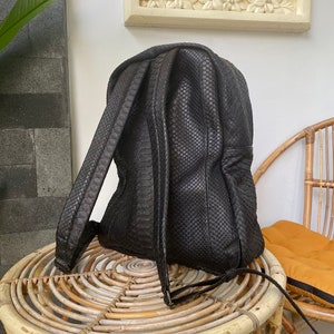 May include: A black leather backpack with a snakeskin pattern. The backpack has a zipper closure and adjustable straps. It is sitting on a wicker table.