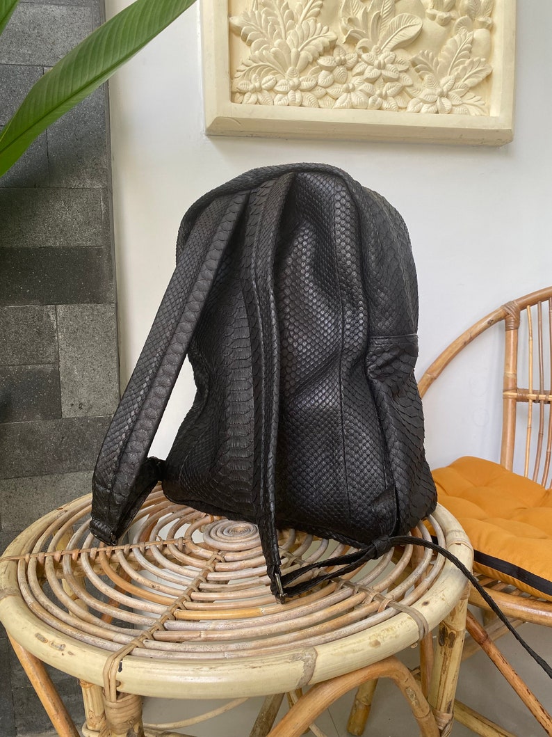 May include: A black leather backpack with a snakeskin pattern. The backpack has a zipper closure and adjustable straps. It is sitting on a wicker table.
