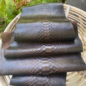 May include: Stack of four dark brown python leather hides with a textured, scaly pattern.