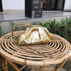 Może przedstawiać: Złota torebka typu kopertówka ze skórzaną fakturą przypominającą skórę węża, z długim paskiem. Torebka leży na okrągłym, plecionym stole.