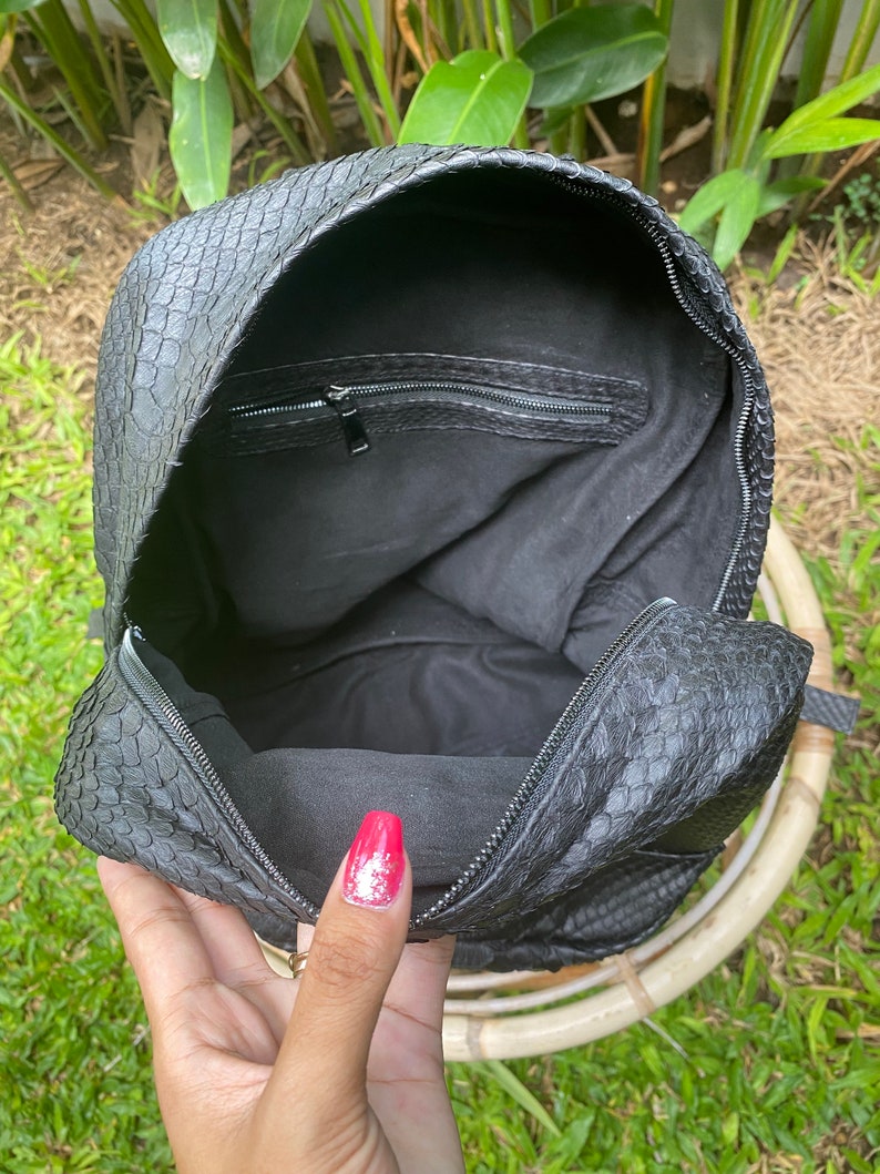 May include: Black leather backpack with a snakeskin pattern. The backpack is open and the inside is lined with black fabric. There is a zippered pocket on the inside of the flap.