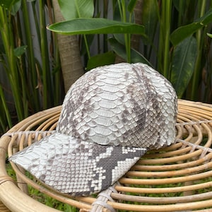 May include: A white and gray snakeskin baseball cap with a curved brim. The cap is made of real snakeskin and has a unique, textured pattern.