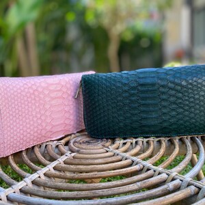 Peut inclure: Deux portefeuilles en cuir, l'un rose et l'autre vert, avec un motif en peau de serpent. Les portefeuilles sont pos&eacute;s sur une surface en osier tress&eacute;.