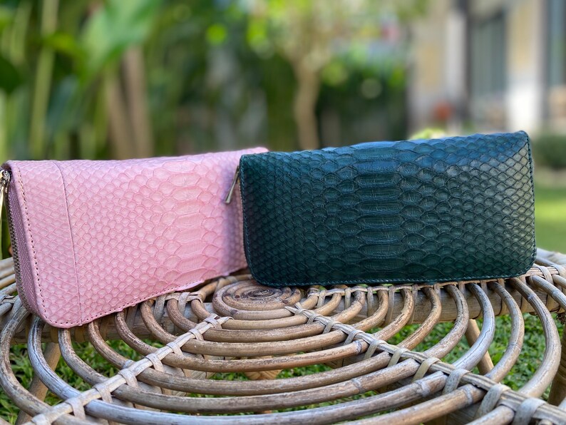 Peut inclure: Deux portefeuilles en cuir, l'un rose et l'autre vert, avec un motif en peau de serpent. Les portefeuilles sont pos&eacute;s sur une surface en osier tress&eacute;.