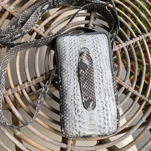 May include: A small, rectangular crossbody bag with a snakeskin pattern in shades of grey and white. The bag has a matching strap and a zip closure. The bag is sitting on a woven wicker chair.
