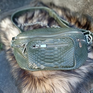 Puede incluir: Una riñonera de piel de serpiente verde con cremalleras plateadas y una correa de cadena plateada. La bolsa está sobre una superficie peluda.