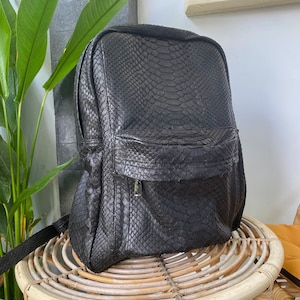 May include: A black leather backpack with a snakeskin pattern. The backpack has a zipped front pocket and adjustable straps.