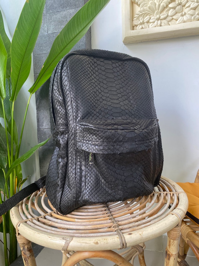 May include: A black leather backpack with a snakeskin pattern. The backpack has a zipped front pocket and adjustable straps.