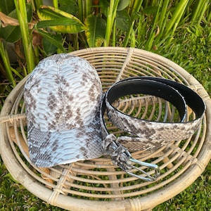 May include: A gray and white snakeskin baseball cap and a matching snakeskin belt with a silver buckle. The cap and belt are on a woven wicker table.